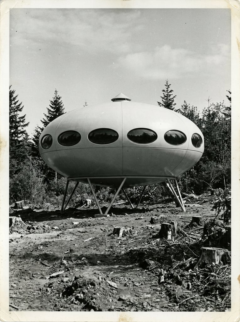 A vintage black and white photograph of a futuristic, saucer-shaped architectural dwelling elevated on angled metal struts in a wooded clearing. The smooth, oblate spheroid structure features a distinct horizontal seam, a continuous band of dark elliptical windows, and a small conical cap on its roof. The building rests in a rugged environment characterized by rough dirt, chopped tree stumps, and scattered evergreen trees beneath a clear sky. The low-angle perspective and high-contrast aesthetic emphasize the striking space-age design against the unrefined landscape.