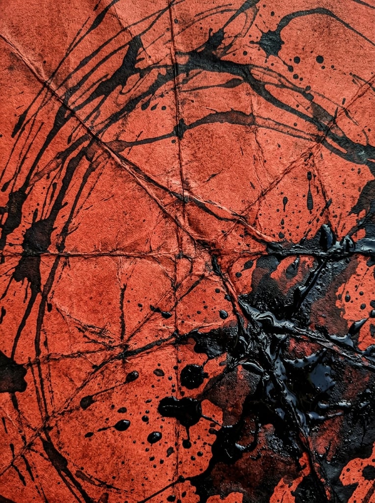 A macro shot of deeply creased rusty red paper covered in erratic, flowing dark ink strokes and heavy splatters. Prominent intersecting fold lines map the distressed surface. Viscous black ink pools and droplets concentrate in the lower right, contrasting sharply with the vivid blood-orange background to create a raw, moody grunge aesthetic.